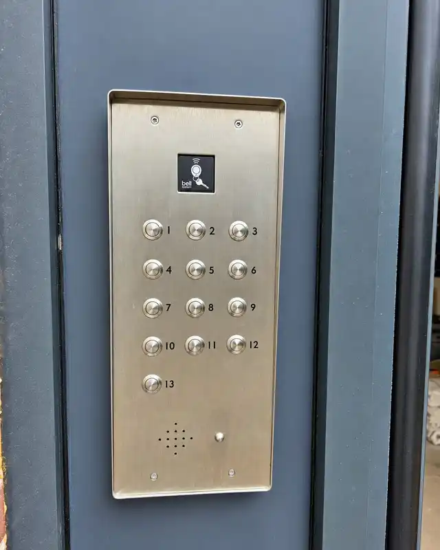 Stainless steel 13-button audio door entry panel with a fob reader at the top, fitted to a dark grey door frame.