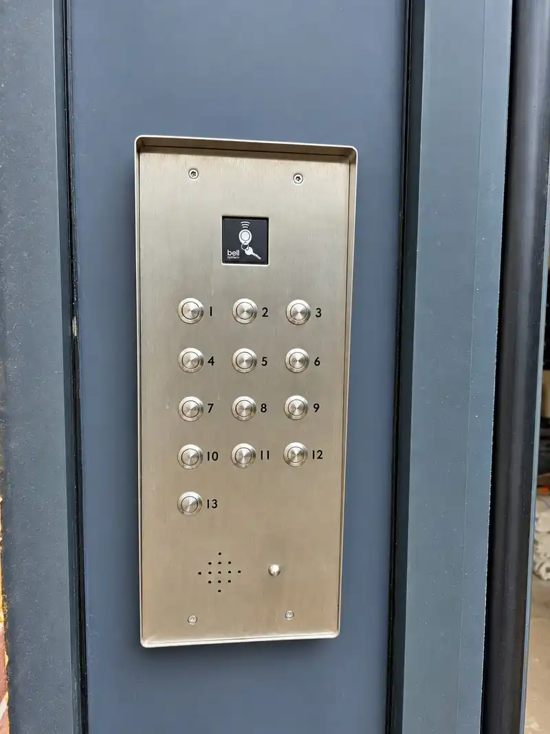 Stainless steel 13-button audio door entry panel with a fob reader at the top, fitted to a dark grey door frame.