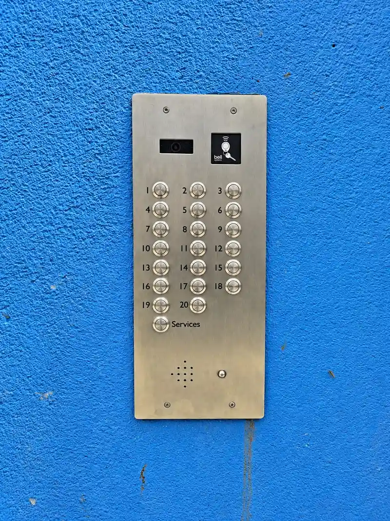 Stainless steel 20-button video door entry panel with a camera and a fob reader, fitted to a blue rendered wall.