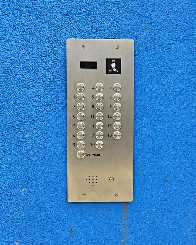Stainless steel 20-button video door entry panel with a camera and a fob reader, fitted to a blue rendered wall.