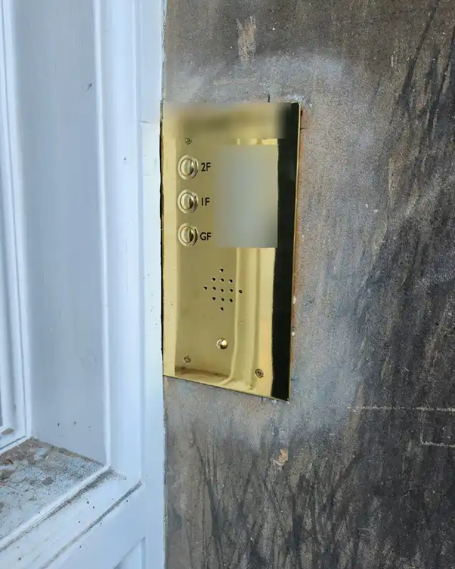 Brass audio door entry panel with three flat buttons and name labels, fitted to a stone wall beside a white door frame.