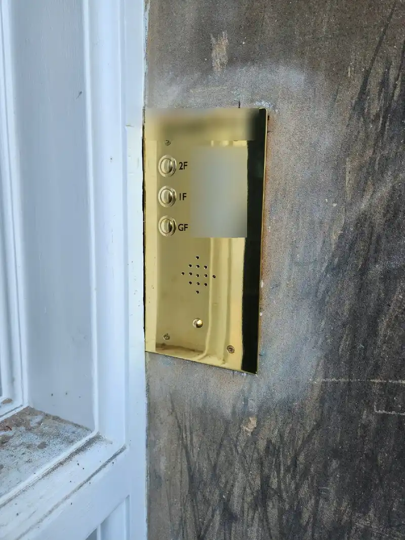 Brass audio door entry panel with three flat buttons and name labels, fitted to a stone wall beside a white door frame.