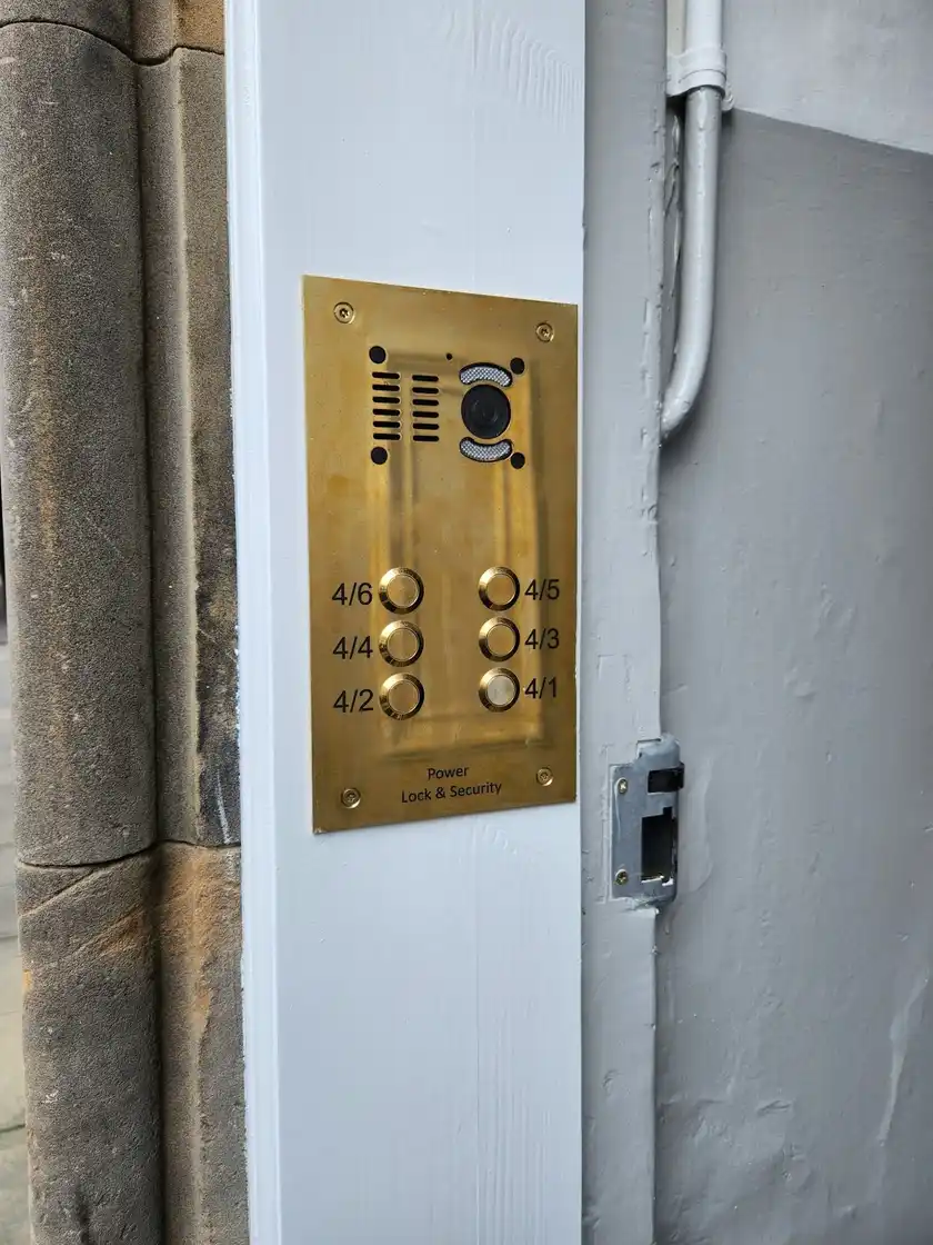 Brass video door entry panel with a camera and six flat buttons, engraved Power Lock & Security, on a white door post.