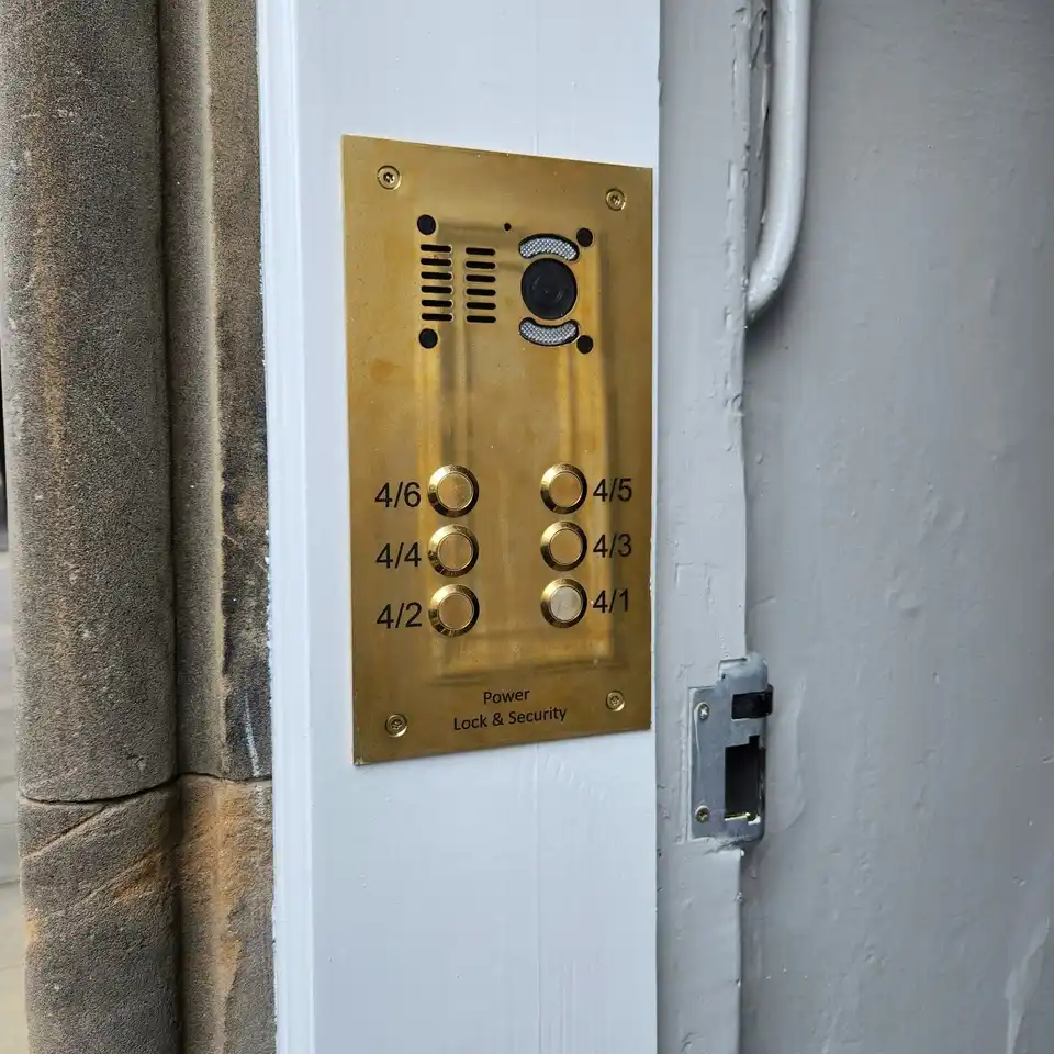 Brass video door entry panel with a camera and six flat buttons, engraved Power Lock & Security, on a white door post.