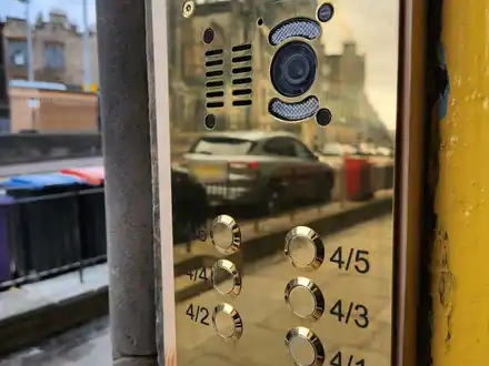 Polished brass video door entry panel with a camera and flat buttons, fitted beside a stair door on an Edinburgh street.