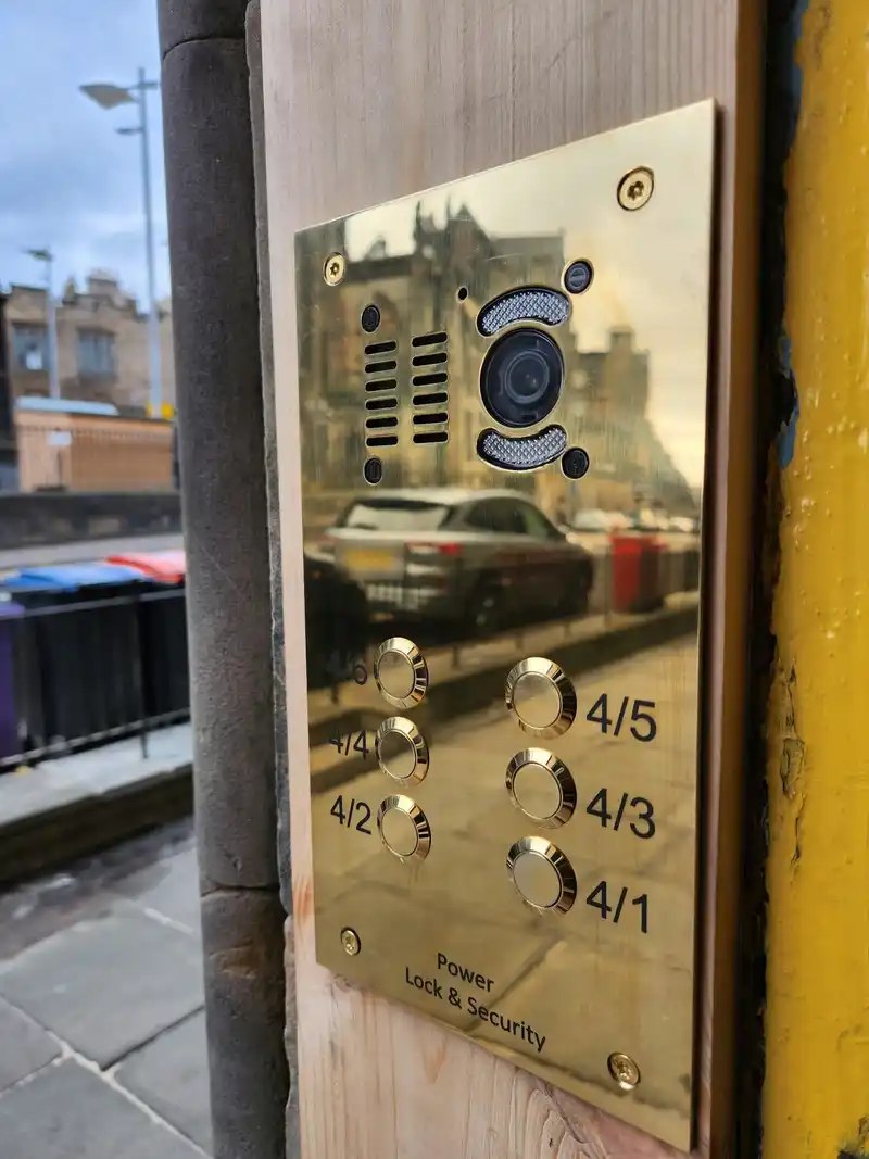 Polished brass video door entry panel with a camera and flat buttons, fitted beside a stair door on an Edinburgh street.