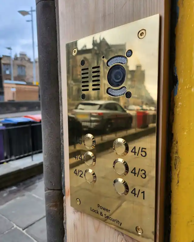 Polished brass video door entry panel with a camera and flat buttons, fitted beside a stair door on an Edinburgh street.