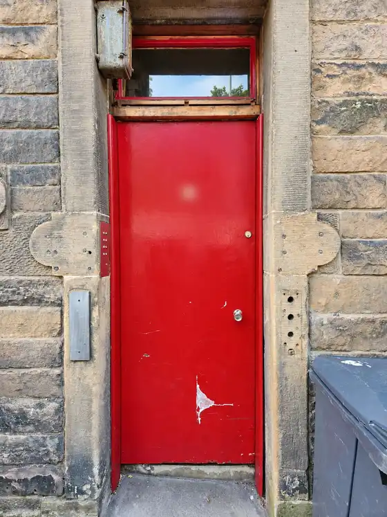 Worn red flat door in a stone tenement doorway, before replacement. House number blurred.