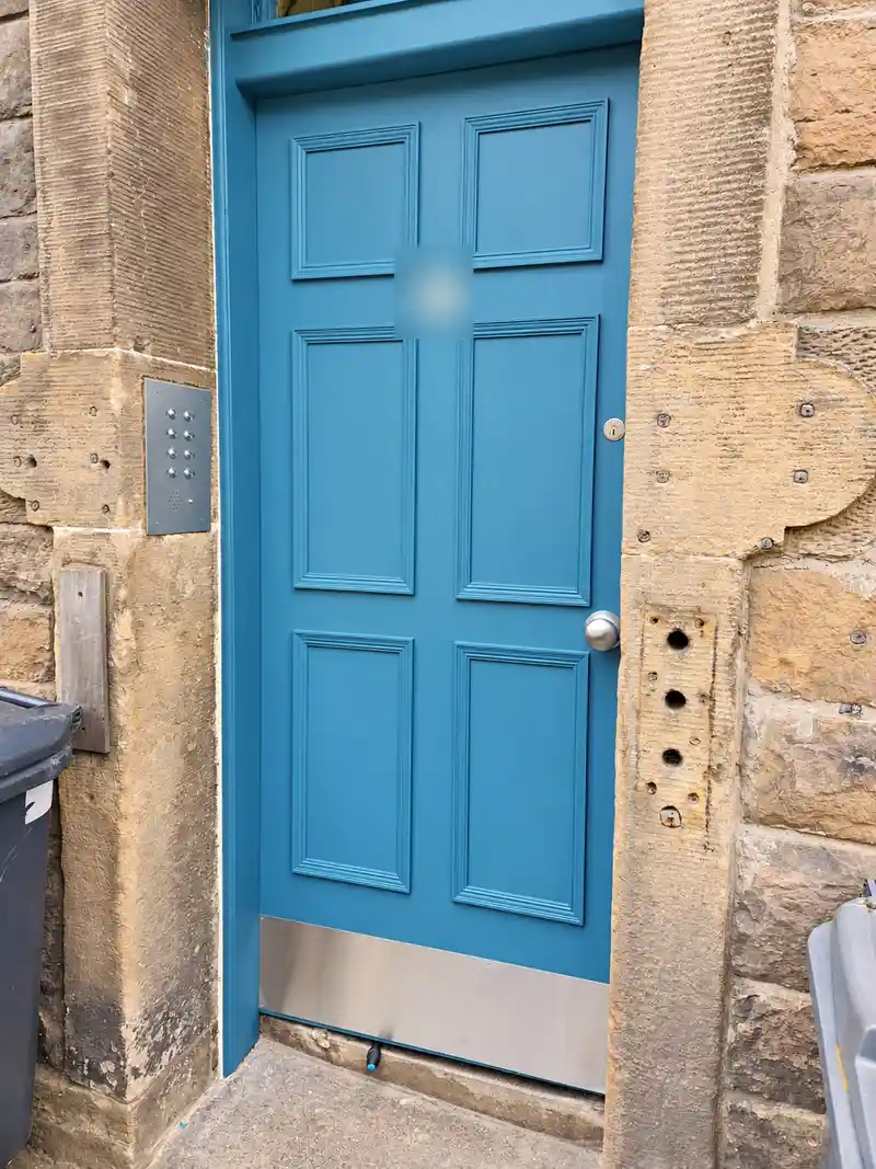 The new six-panel door painted teal-blue, with a steel kick plate and an entry panel on the wall to the left.