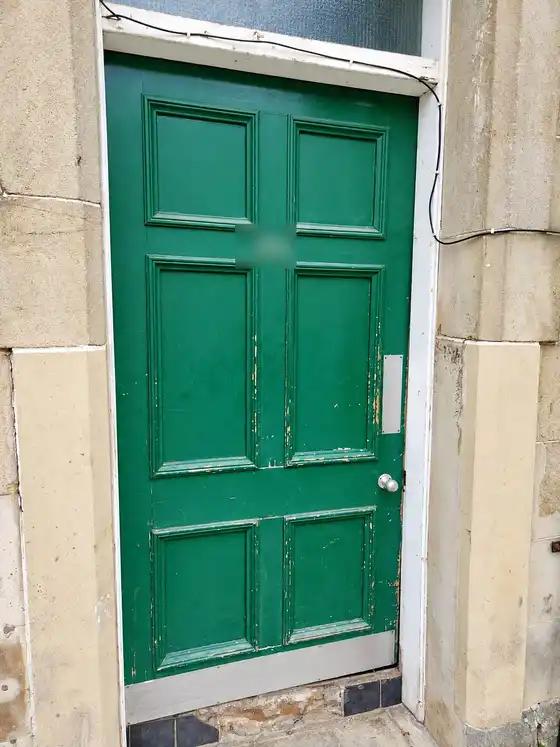 Old green panelled door with flaking paint in a sandstone surround. House number blurred.