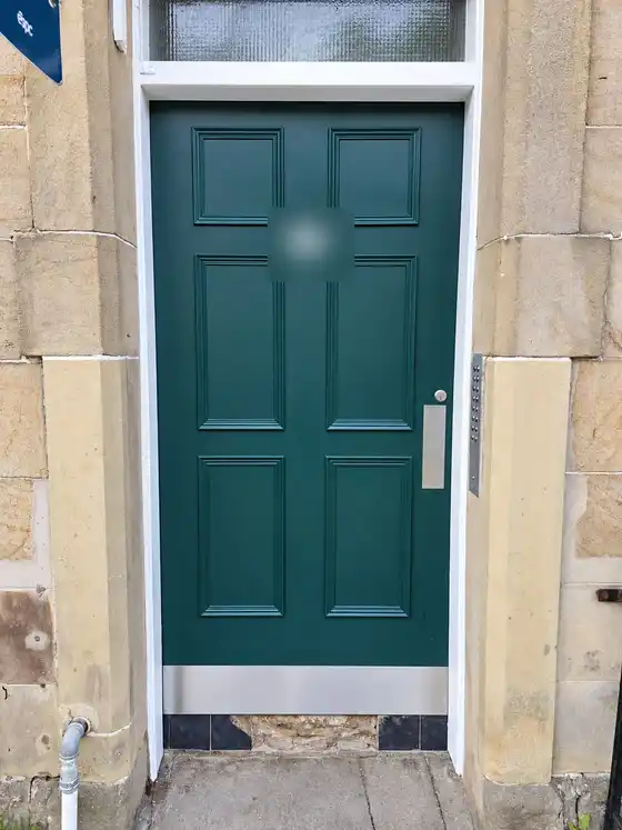 New six-panel stair door painted dark green, with a steel kick plate, in a sandstone surround with a fanlight above. House number blurred.