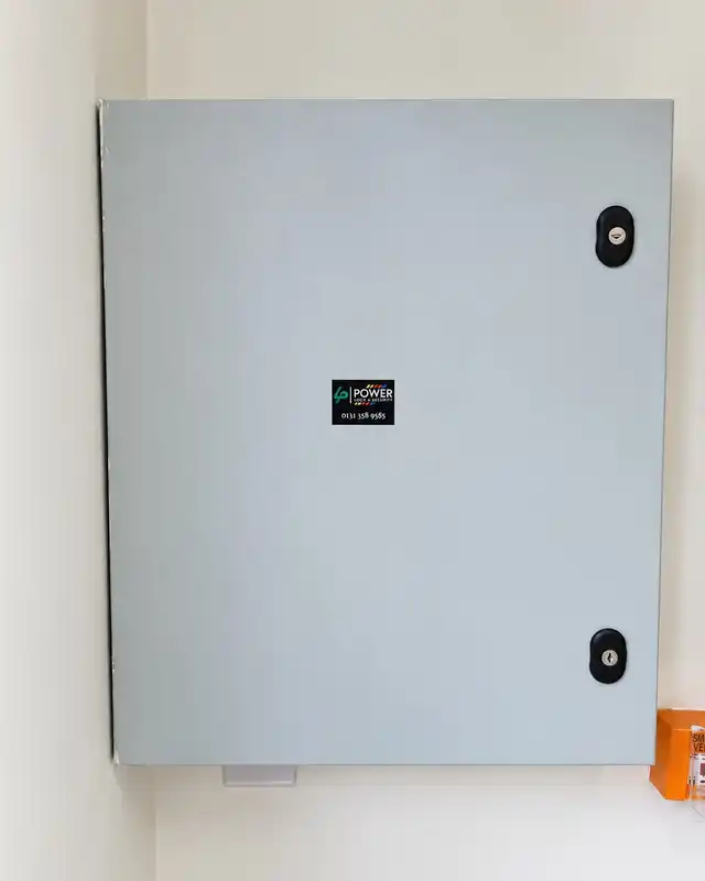 Grey steel equipment enclosure on a white wall with a small Power Lock & Security sticker, and an orange break-glass unit below.