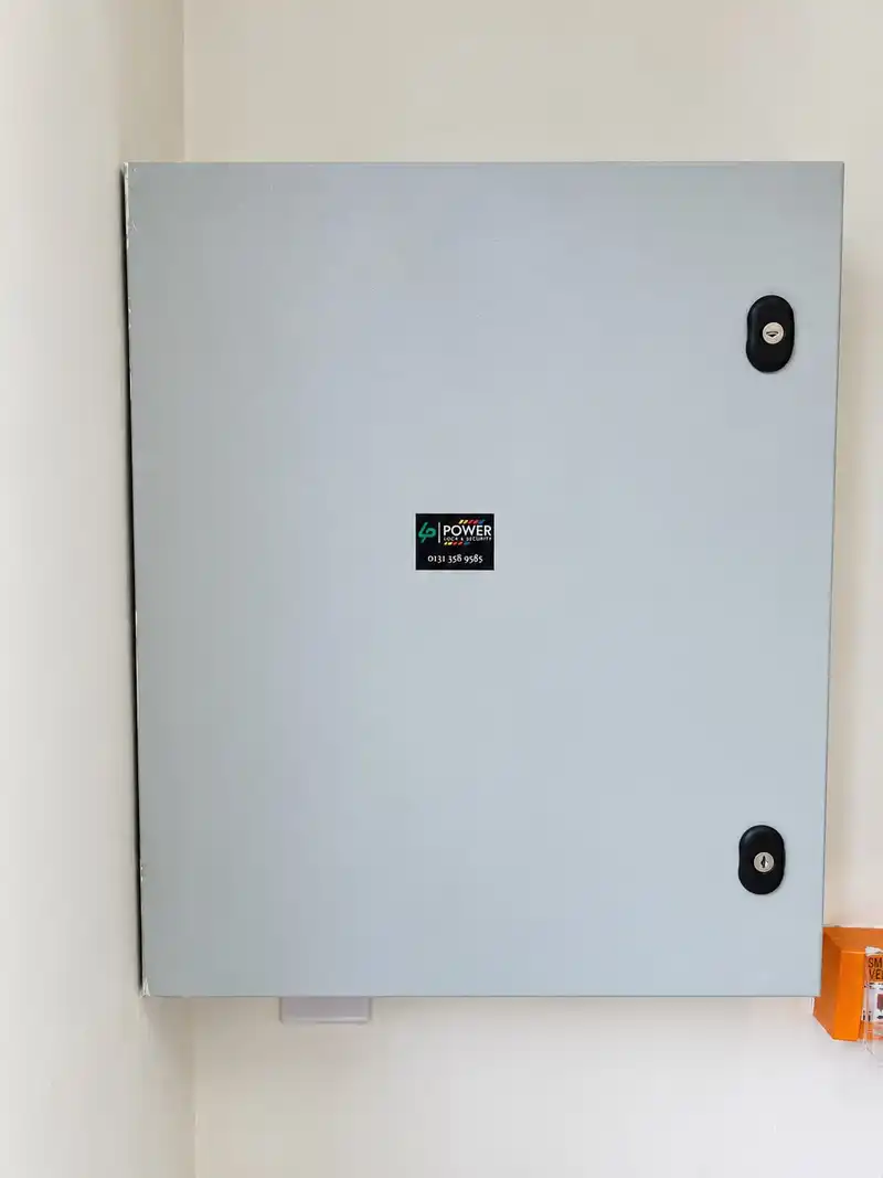 Grey steel equipment enclosure on a white wall with a small Power Lock & Security sticker, and an orange break-glass unit below.