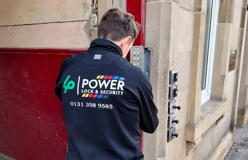 Engineer in a Power Lock & Security jacket working on a door entry panel beside a sandstone doorway.