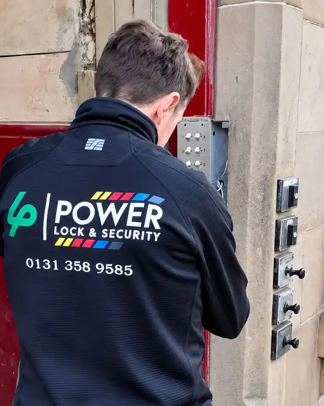 Engineer in a Power Lock & Security jacket working on a door entry panel beside a sandstone doorway.