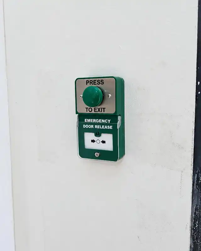 Green "press to exit" button mounted above a green emergency door release break-glass unit on a white wall.