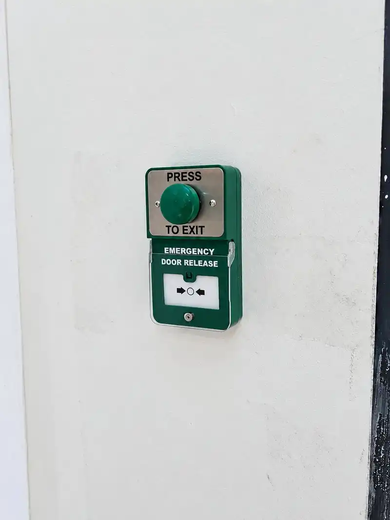 Green "press to exit" button mounted above a green emergency door release break-glass unit on a white wall.