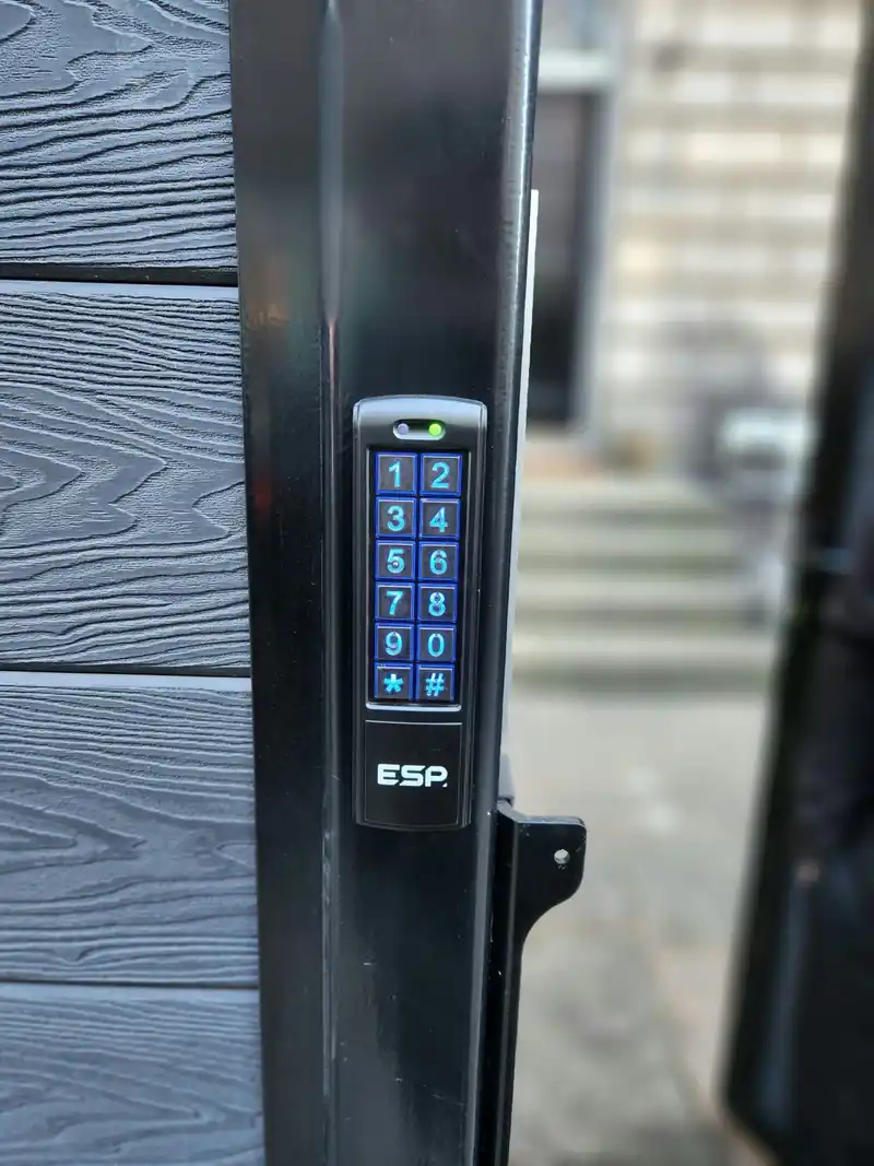 Illuminated blue keypad on the black frame of a grey garden gate.