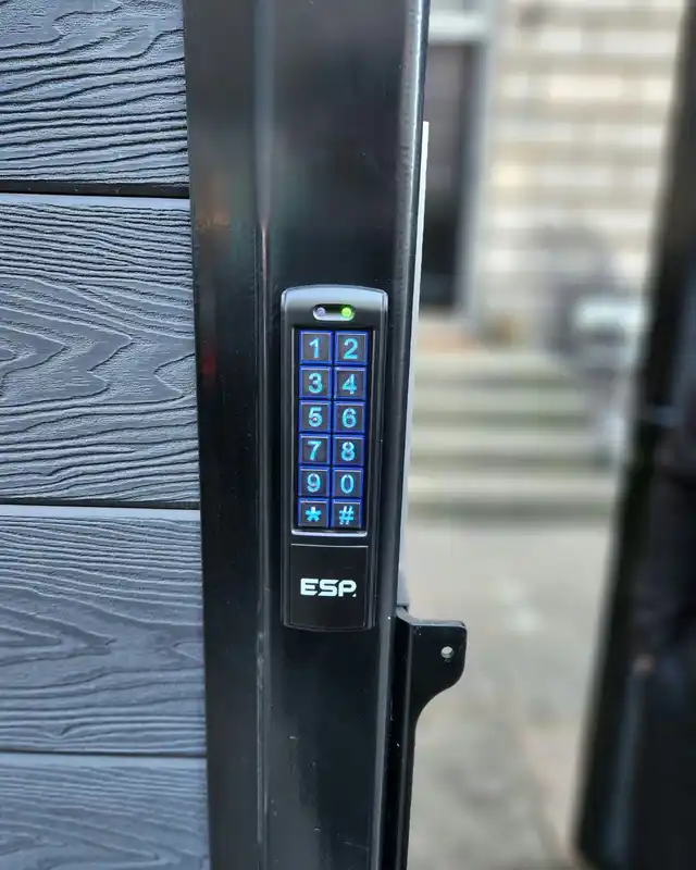 Illuminated blue keypad on the black frame of a grey garden gate.