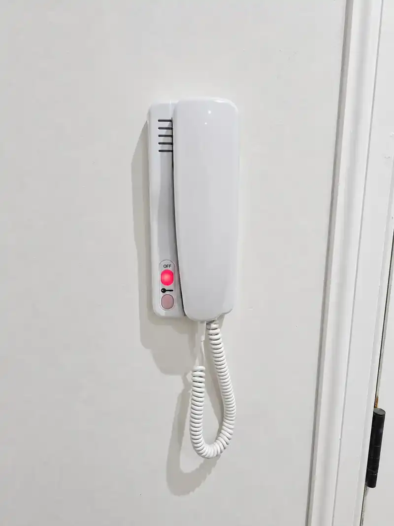 White wall-mounted door entry handset with a coiled cord and a red indicator light.