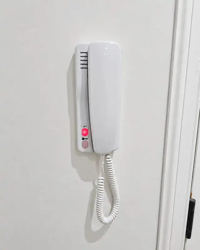 White wall-mounted door entry handset with a coiled cord and a red indicator light.