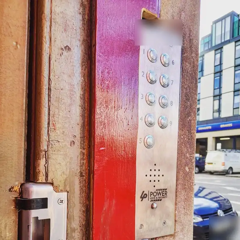 Stainless steel door entry panel with ten call buttons and the Power Lock & Security logo, beside a red painted door frame with an electric lock keep below.
