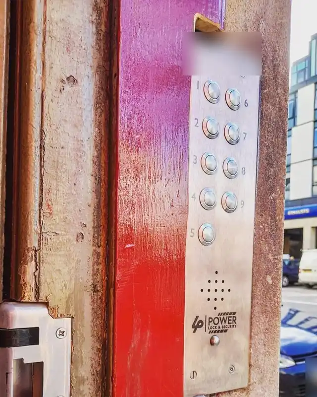 Stainless steel door entry panel with ten call buttons and the Power Lock & Security logo, beside a red painted door frame with an electric lock keep below.