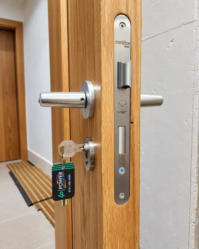 Satin steel lever handle and mortice sashlock on an oak-veneer door, with a corridor behind.