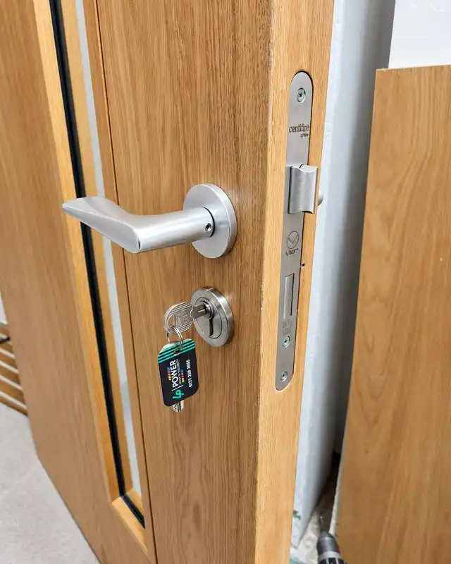 Edge of an oak-veneer door with a lever handle, a euro cylinder with a key in it, and a mortice sashlock forend marked Certifire.