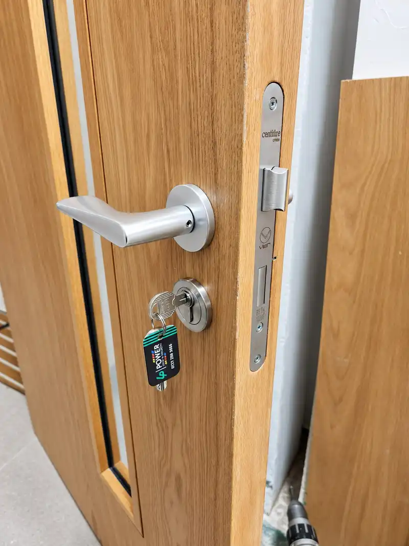Edge of an oak-veneer door with a lever handle, a euro cylinder with a key in it, and a mortice sashlock forend marked Certifire.