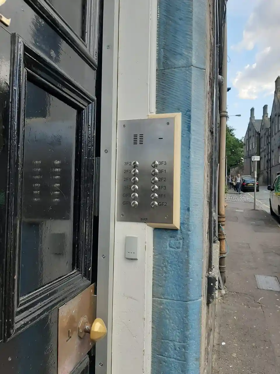 Black tenement front door in a stone entrance with a stainless entry panel labelled by flat position.