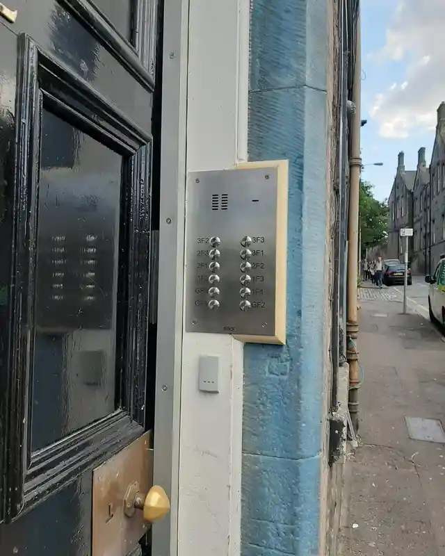 Black tenement front door in a stone entrance with a stainless entry panel labelled by flat position.